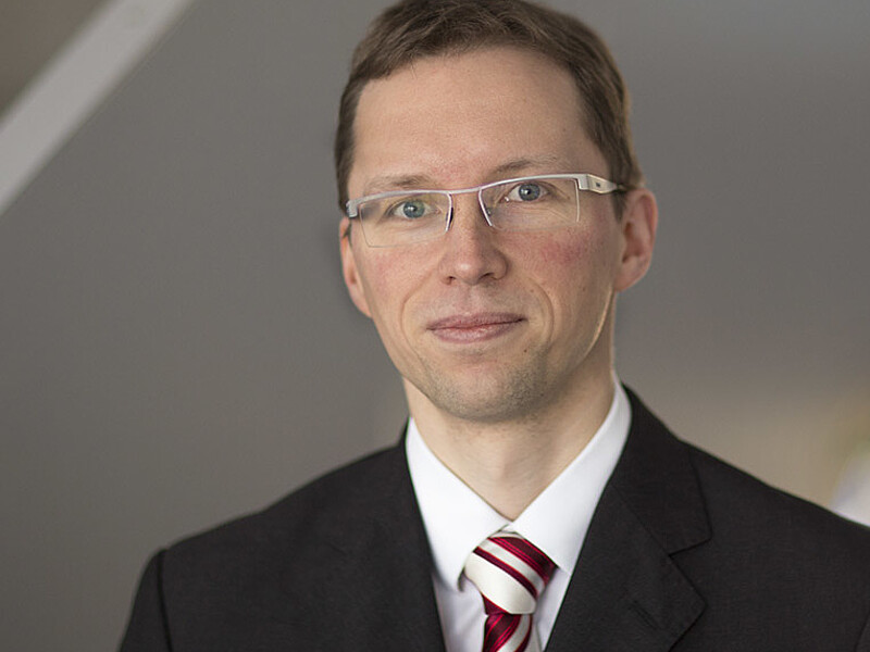 Stefan Kornhuber im Anzug mit Krawatte und Brille lächelt in die Kamera.